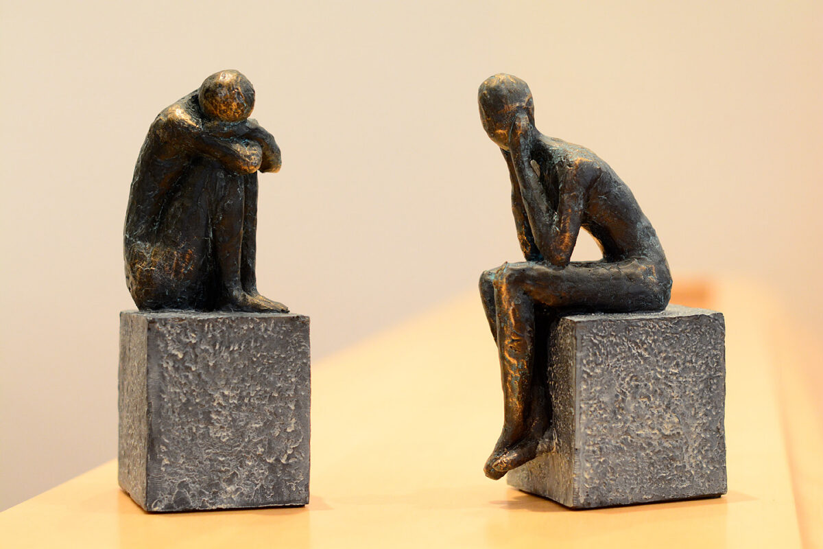 Zwei kleine Bronzefiguren auf Steinblöcken, die nachdenklich und in Trauerhaltung sitzen.